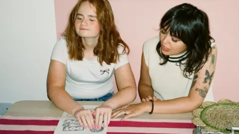 Andy Jude-Williamson Evie Roberts on the left is sitting at a table with Natasha Molyneux-Smith to her right. Evie has medium length wavy red hair and Ms Molyneux-Smith has medium length black hair and a fringe. Evie has her hands to a piece of paper reading braille and Ms Molyneux-Smith is reaching over to see it.