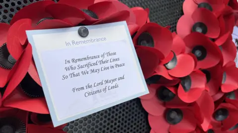 A poppy wreath with a message pinned on it. The message reads: "In Remembrance of those who sacrificed their lives so that we may live in peace. From the Lord Mayor and Citizens of Leeds."