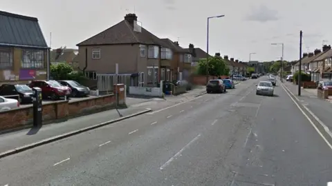 Google Whippendell Road, with some traffic coming towards the camera. There is a parking machine on the left-hand pavement. Houses line both sides of the street. There are dotted lines on the left side marking parking spaces, and a single yellow line on the right.