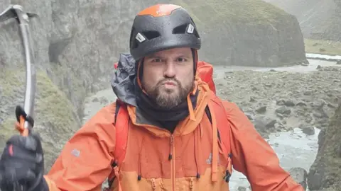 Lost Earth Adventures A climber wearing an orange jacket, helmet, and harness ascends a steep rocky waterfall while holding an ice axe. Mist hangs below, with dark rock walls rising sharply on either side and a stream running through the base.