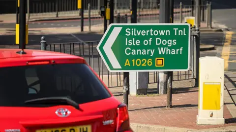 A green road sign pointing left toward "Silvertown Tnl," "Isle of Dogs," and "Canary Wharf" on the A1026. A red car and a white bollard are in the foreground.