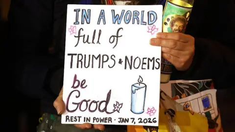 East Bay Times via Getty Images A close up shot of a woman holding a decorated sign that says 'in a world full of Trumps and Noems, be good. Rest in power. Jan 7, 2026'. 