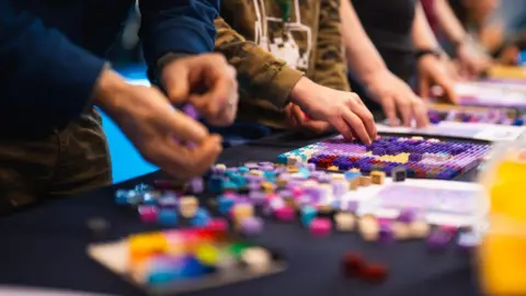 A number of unidentified people can be seen lined behind a table playing with various Lego kits.