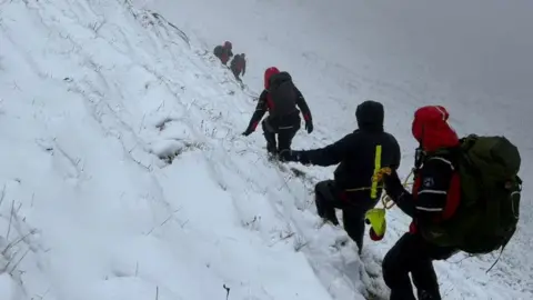 Five people in waterproof gear walking in line across a steep snow covered hill