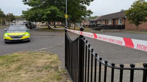 A police car which is yellow and blue in colour is parked to the left of a police cordon tape. Police is written on the side and front of the car in blue letters. To the right of the picture are houses and to the fore are black railings with the police tape across them.