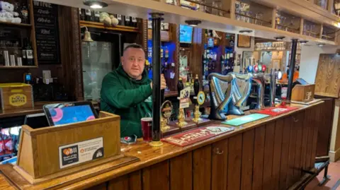 A man in a green hoodie standing behind a bar and pulling a pint. He is looking at the camera. 