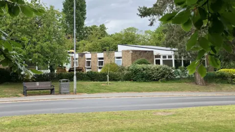 A brick one-storey building with windows and surrounded by trees. There is a road, a pavement, a bench, a waste bin and a lamp post in front.