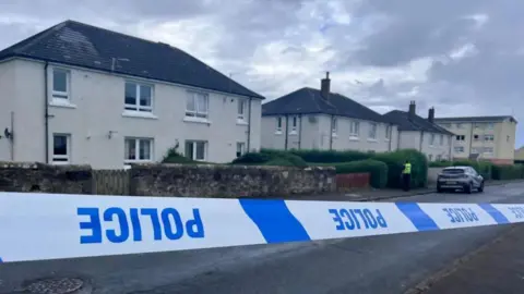 Blue and white police tape which is upside down in front of a row of cream two-storey buildings. A police officer is visible in a distance.