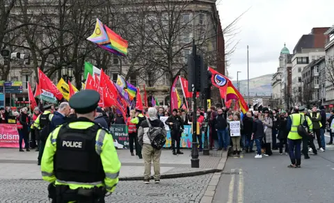 Pacemaker Police office in foreground, standing facing a large crowd of protesters holding colourful trade union and pride flags, on Donegall Square in Belfast with large buildings in background.