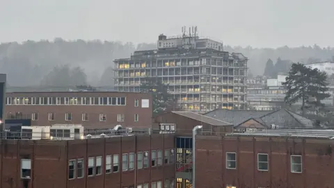 A view of Wycombe Hospital building on an overcast day, with lights visible in the windows and trees and other buildings in the background.