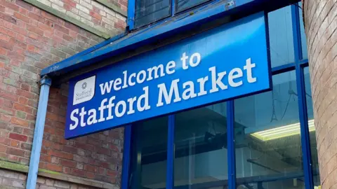 Blue signage with white letter reading 'welcome to Stafford Market' with the logo of Stafford Borough Council. Behind the sign there is a window and a brick wall either side.