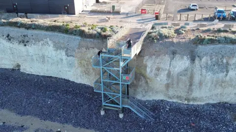 A metal staircase leading down from a cliff top car park onto a shingle and sand beach.