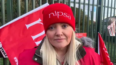 BBC A woman with long blonde hair wears a red jacket with a green coat underneath as well as a red beanie hat with the letter NIPSA on it, in white font. Behind her is green metal fencing which has red flags resting on it. Green bushes and a blue sky can be seen through the fence. A black poster with white fists reads 'Strike' and 'Stailc' in black writing.