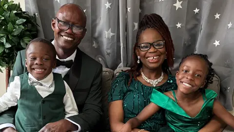 A woman with glasses, a necklace, a long green dress, and a man with glasses and a green tuxedo, sit on a light grey sofa with their son, who is wearing a green waistcoat and suit, and daughter, who is wearing a green dress. All four of them are smiling. A plant is behind the man's right shoulder. Curtains behind them are grey and have white stars on them.