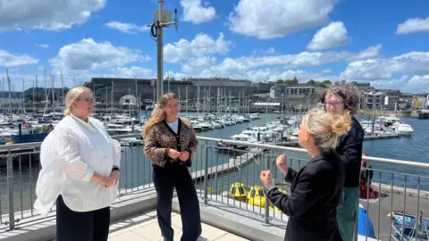 on the left and stood next to Rebecca Hegedus. They are stood opposite their interpreters. They are all smiling at one another and are stood on the balcony with a view of Queen's Anne Battery where there are docked boats. 
