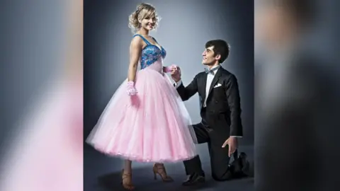 Tina O'Brien smiles at the camera, wearing her pink and blue ballroom dress, standing next to her dance partner Jared Murillo, who is wearing a dark tailcoated outfit with grey bow tie.