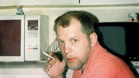PA Media Michael Stone, holding a cigarette in his right hand and looking towards the camera. He has heavy stubble and is wearing a peach-coloured top. A microwave oven can be seen behind him.