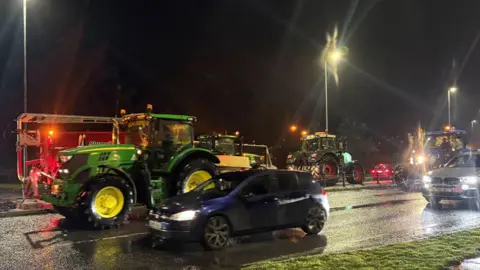 A number of tractors and cars driving down a road in convoy. It is dark and the scene is lit up by street lights. 