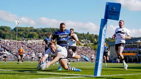 PA Media Bristol's Max Malins scores their first try during the Gallagher Premiership match at The Recreation Ground, Bath. He is diving over the line holding the ball while a packed stand is visible in the background, as are the hills of the city beyond