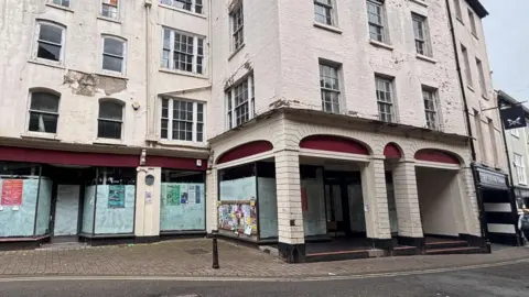 A large white building with a front with arches and pillars and some of the brickwork appears to be cracked and falling off. There are boarded up shops on the ground floor.