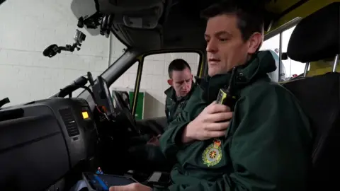 Two male paramedics are sitting in the front of an ambulance. One is speaking into a radio. They are wearing green uniforms.