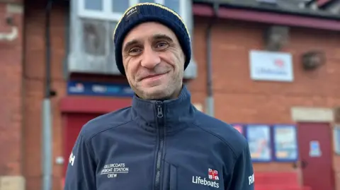 BBC Carl Taylor smiles at the camera while standing in front of the red brick lifeboat station. He is wearing a navy jacket and hat with RNLI logos, and has faint stubble.