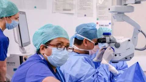 South Tyneside and Sunderland NHS Foundation Trust A cataract operation taking place at Sunderland Eye Infirmary. Four medical staff with face masks, scrubs and gloves have surrounded an operating table. One of the clinicians is looking through a machine.
