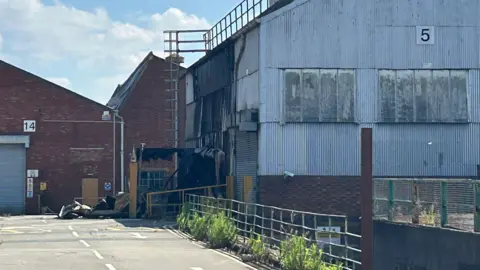 A red brick building covered in metal sheeting has scorch and burn marks across one side. 