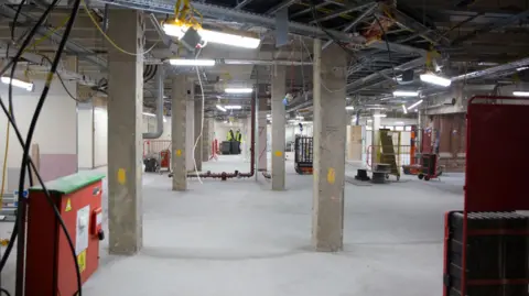 Image of the inside of a building under construction. The building has an open ceiling showing all the ductwork, a concrete floor, a number of concrete pillars and strip lighting. In the foreground is a red metal power station.