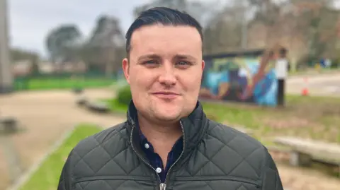 Dark-haired man wearing a grey jacket and blue shirt looking at the camera, with a blurred school behind him