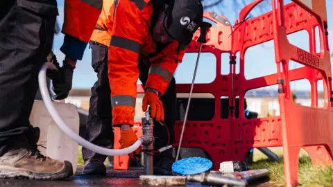 Origin Tech A man wearing a hi vis orange jacket and black cap with the words 'no dig' on it. He is bending down and holding machinery with a long white tube in the ground