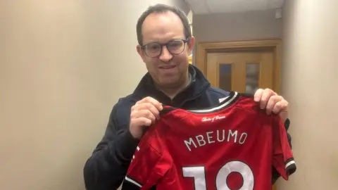 A man holding a Man Utd shirt that says Mbeumo 19. He has dark hair and glasses.