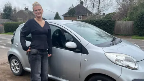 Young woman wearing a black v neck long sleeved top standing by a silver car and smiling. 