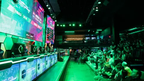 MEWC A view of audience members sat in a seating area to the right and the MEWC final competitors area to the left. The arena is filled with screens and is bathed in a green light.
