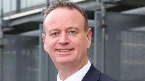 Stephen Kelly is wearing a black suit and white shirt. He has short brown hair and blue eyes. He is standing in the campus in Londonderry. 