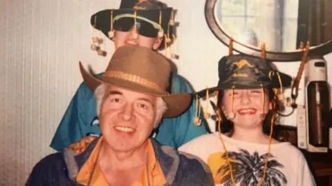 An old photo of two children and their grandad wearing Australian cork hats. A man with grey hair is in the middle wearing a stripy apron and a brown hat.
