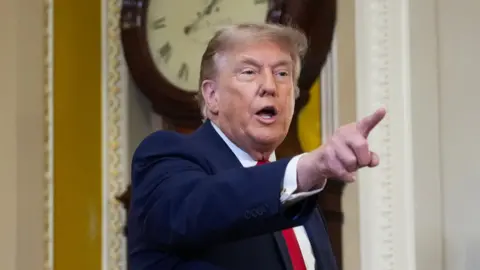Donald Trump wearing a navy suit and red tie, points his finger to the right side of the frame.