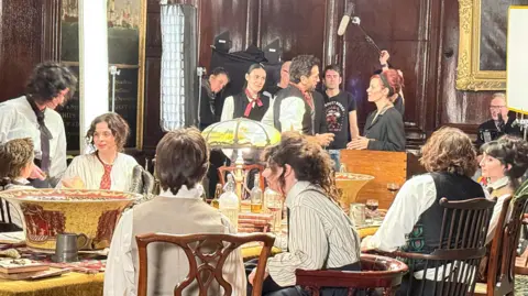 A behind-the-scenes shot from Night and Day which shows a group of actors in costumes, some who are sitting around a table and some who are standing up. The photo also shows crew members with a camera and holding an audio boom to record what is happening 