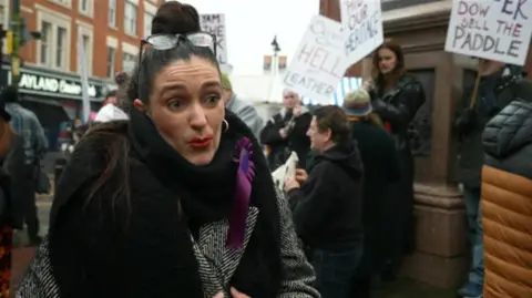 A woman with black hair pulled into the bun looks at the camera as she talks. Behind her are groups of protestors with campaign signs, standing in Walsall town centre. She wears a black scarf, and black and white striped coat.