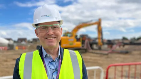 Owen Sennitt/BBC Kieran Healy standing in front of metal fencing at the construction area at Firside Junior School. He is looking directly at the camera and smiling. He is wearing a blue shirt with a blazer over the top and a yellow fluorescent  