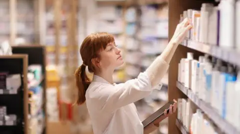 Pharmacist looking at shelves of medication. She wears a white lab coat.