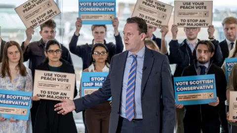 PA Media A person in a suit stands in front of a group holding signs that read ‘Vote Scottish Conservative,’ ‘Use Your Peach Vote,’ and ‘To Stop an SNP Majority,’ with the group arranged indoors in front of large windows