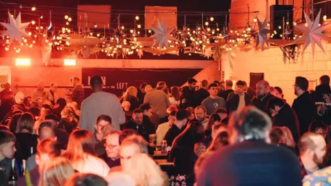 Bustler Market Derby A sea of people sat at wooden tables. Some pint glasses are visible. The image is taken at night time. 