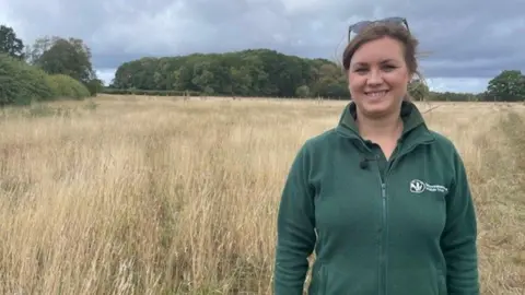 A woman has sunglasses on top of her dark hair and she is wearing a green top. She is standing in a field and trees are in the distance.