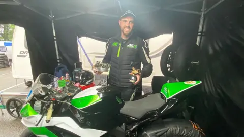 Aran Sadler stands behind a green and white motorcycle holding two trophies. He has black hair and beard and is wearing black biking gear and a baseball hat