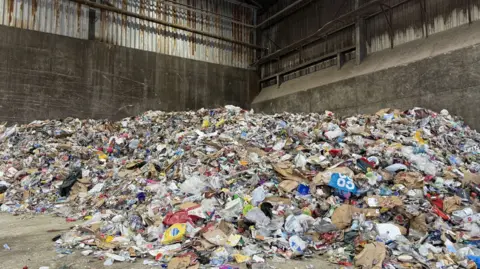 A huge pile of recycling, with waste items of many colours, in a concrete warehouse.