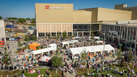 A lively outdoor event taking place in the square. The area is bustling with people who are gathered around white canopy tents, which appear to be hosting stalls or activities. The square is decorated with colourful bunting. Many attendees are seated on the grass and paved areas, enjoying the sunny weather.
In the background, there are large commercial buildings, including a prominent Cineworld cinema with its logo clearly visible on the beige facade. To the right, there is a TK Maxx store, indicating that this is a shopping and entertainment district. The sky is clear and bright blue.