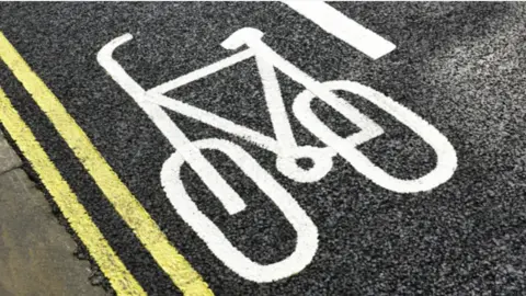 A cycle path with a white painted bike symbol on black tarmac, with two yellow lines to the left. 