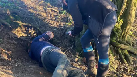 Ben Gray Two people are looking at a hole on a forest floor. They are dressed in outdoor clothes.
