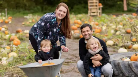 Zoe Rhiane Photography A woman with long brown hair, wearing a black shirt, pants, holds a young boy with a wheelbarrow. A man with dark hair is crouched down next to her, holding a young boy with long, blonde hair. A field is behind them with pumpkins.
